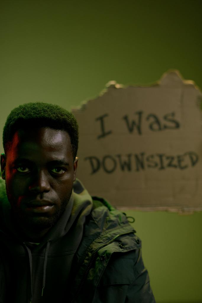 Photo of a Black man looking sad with a cardboard cutout written behind him with the words "I was downsized" on it.